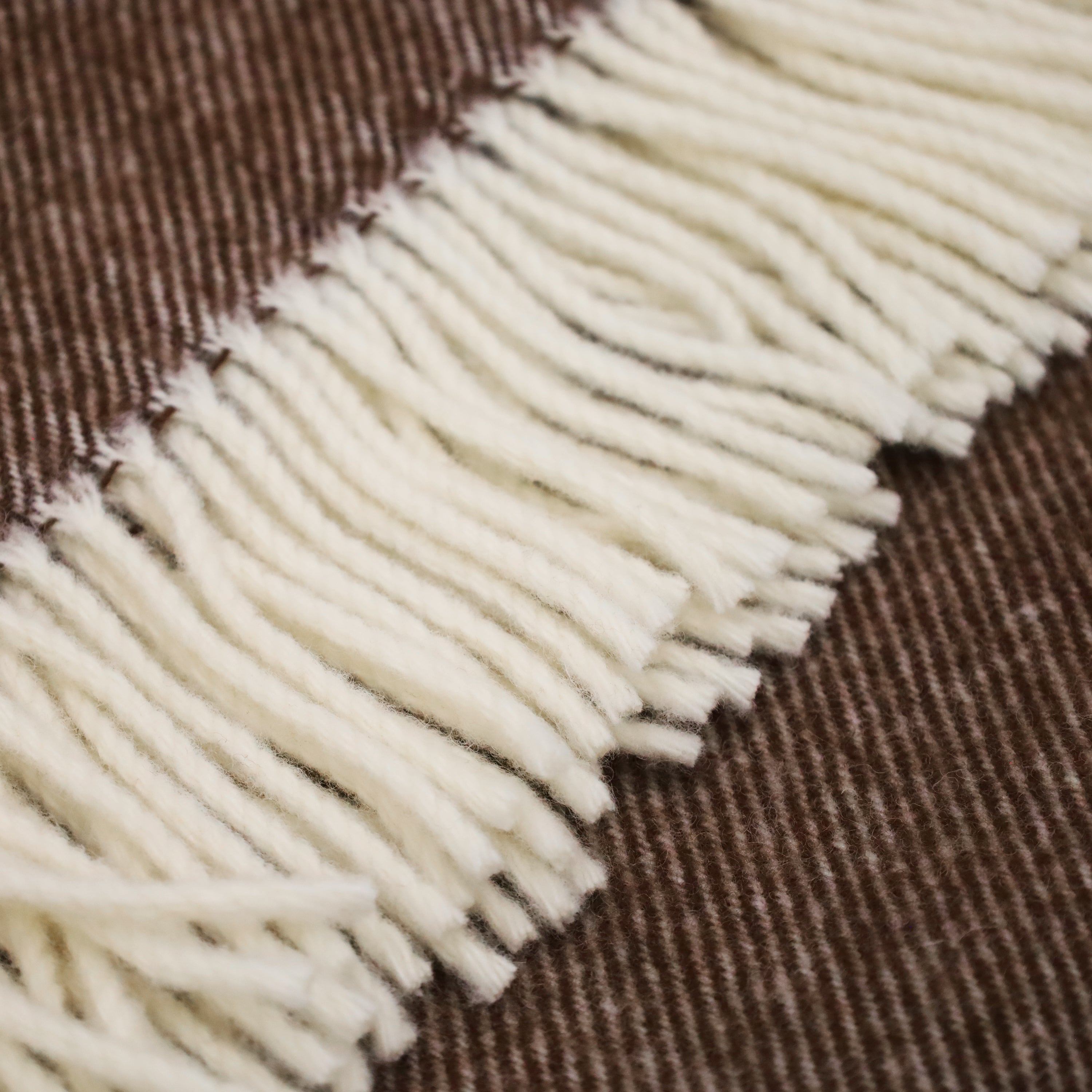 Gros plan dun jeté en laine mérinos plié de Humble Nature. Ce textile en laine marron et crème de NUTSHELL présente un motif rayé et une section crème avec des franges lâches, créant une texture et un contraste sur le fond marron, incarnant une élégance raffinée.