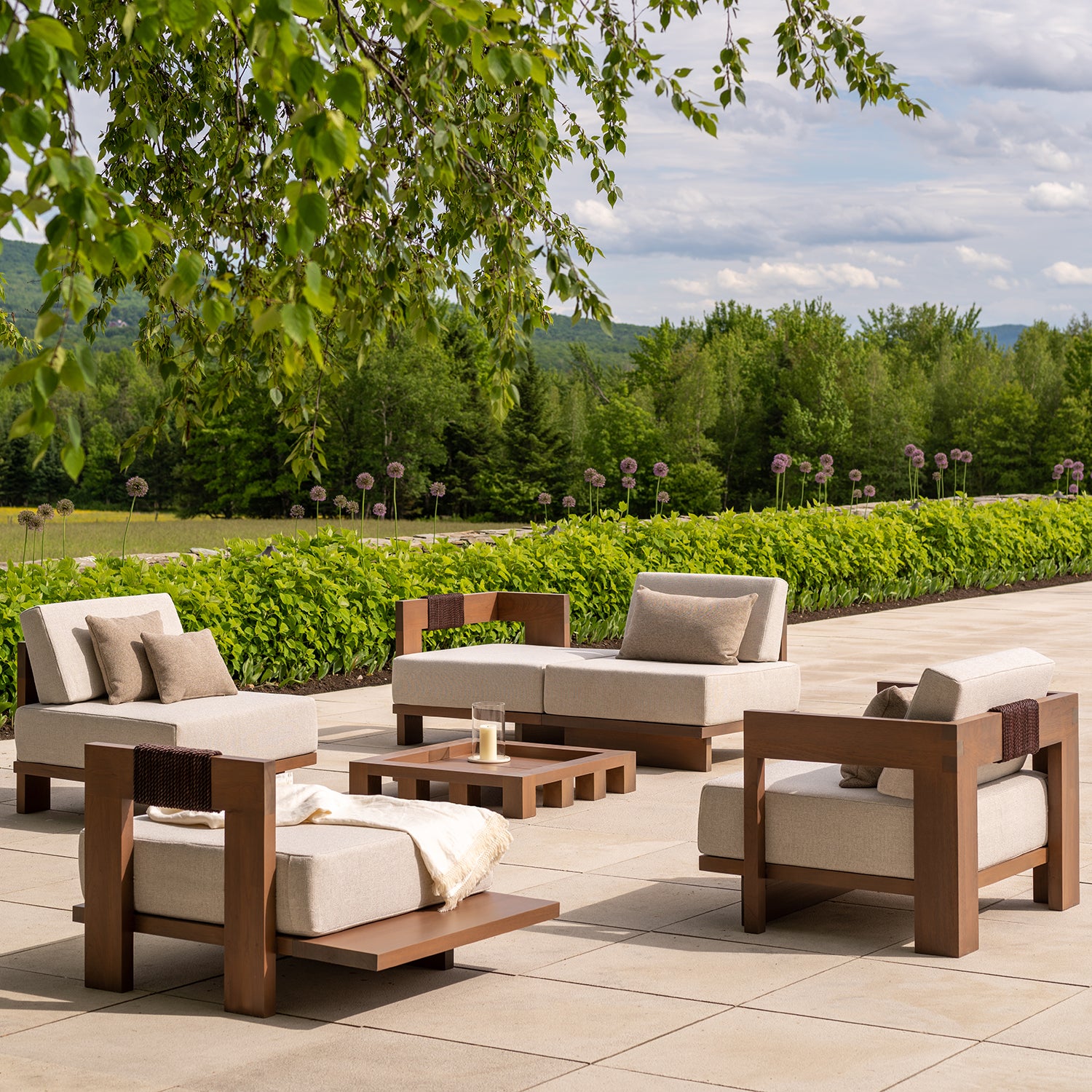 Un ensemble de patio extérieur comprend des chaises en bois coussinées fabriquées à partir de bois écoresponsable et une Table à café | Les Ensembliers de Richard Ouellette et Maxime Vandal. Une couverture et un verre sont posés sur la table, avec des arbustes verts luxuriants, des arbres et un ciel nuageux offrant une toile de fond élégante.