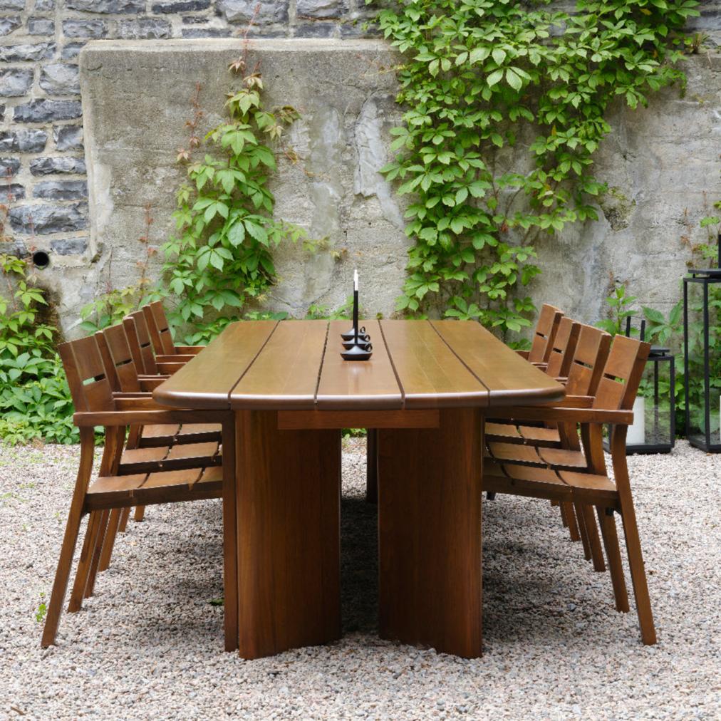La table Horizon 72 de la gamme Roussette de Humble Nature, fabriquée en bois écoresponsable, accompagnée de six chaises assorties, repose sur du gravier. Une bougie dans un support en métal orne le centre. Un mur de pierre avec du lierre vert rehausse le charme de cette collection durable.