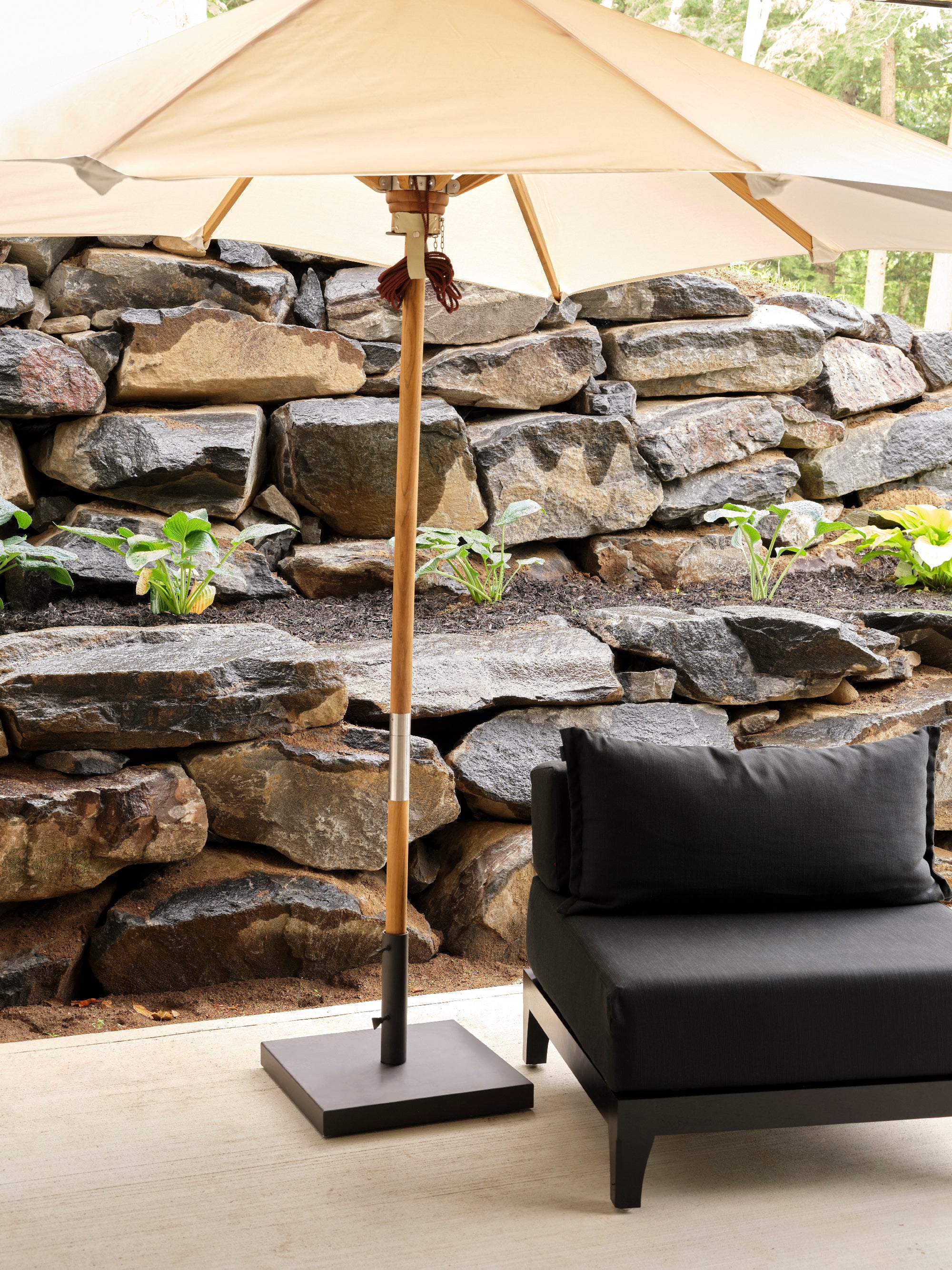 Une scène de patio chaleureuse met en valeur une chaise noire rembourrée placée sous le parasol rectangulaire en bois de teck Humble Nature | Slate. Le mur de pierre en arrière-plan, orné de petites plantes vertes, rehausse le charme du design contemporain, créant un extérieur accueillant et paisible.