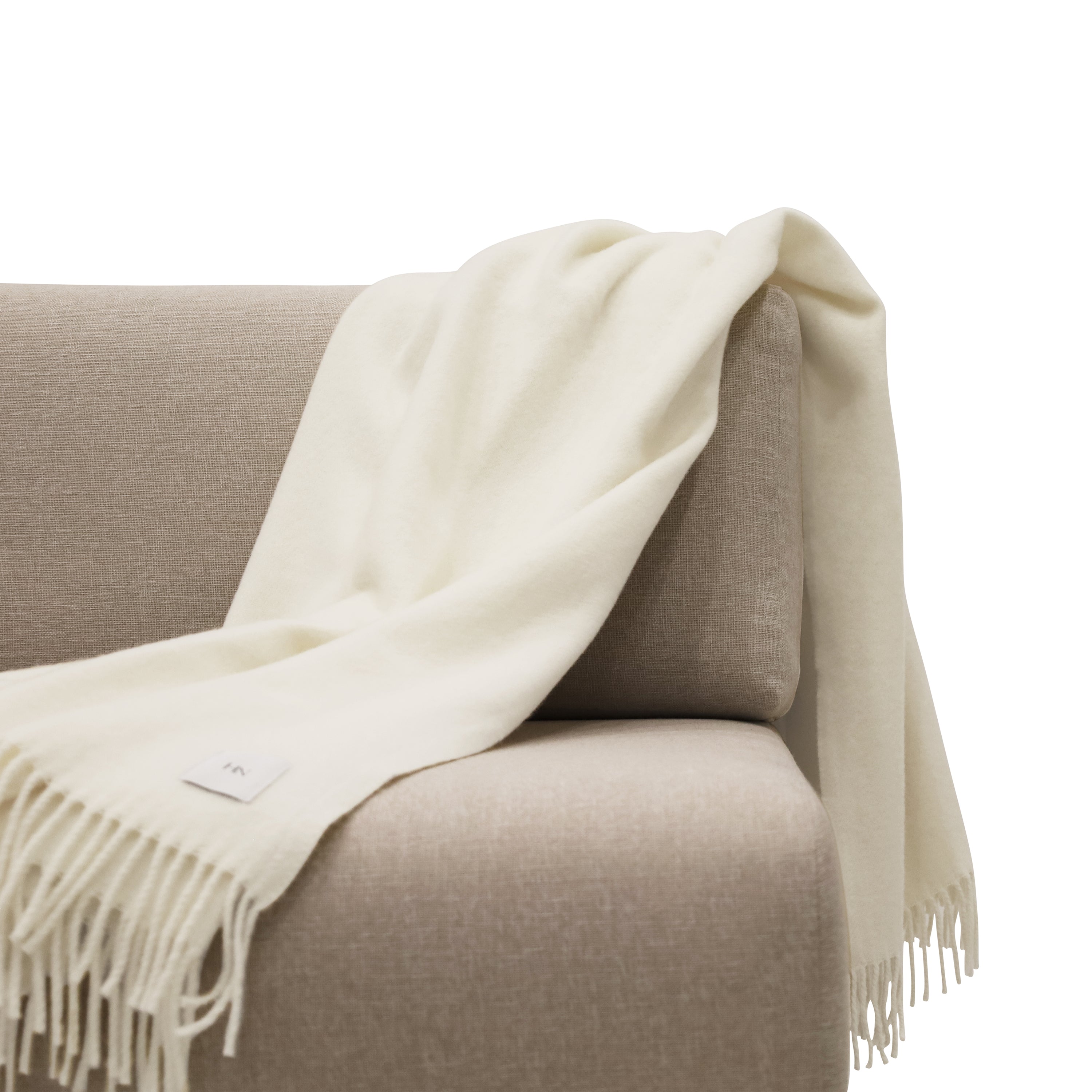 Le jeté en laine mérinos │ SHADE de Humble Nature, une couverture à franges de couleur crème, est élégamment drapé sur un canapé beige sur fond blanc. Fabriqué en laine mérinos fine et doté dune petite étiquette avec des initiales, il ajoute une touche de sophistication à ce décor chaleureux.