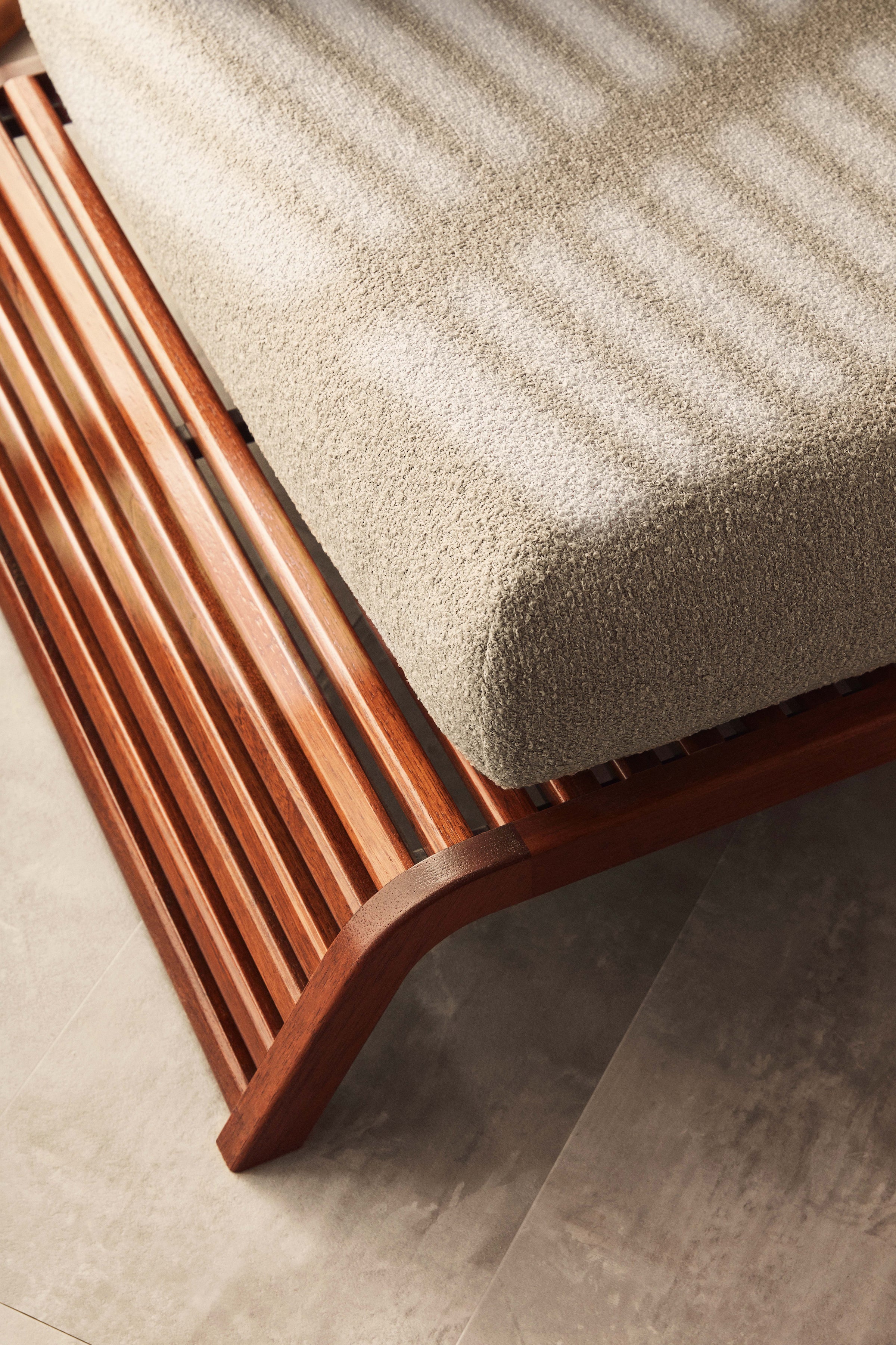 Un gros plan du canapé double extérieur Horizon de Humble Nature montre sa structure métallique à lattes sous un matelas texturé de couleur claire. La lumière du soleil projette des ombres rayées et le sol en carreaux de pierre se trouve en dessous.