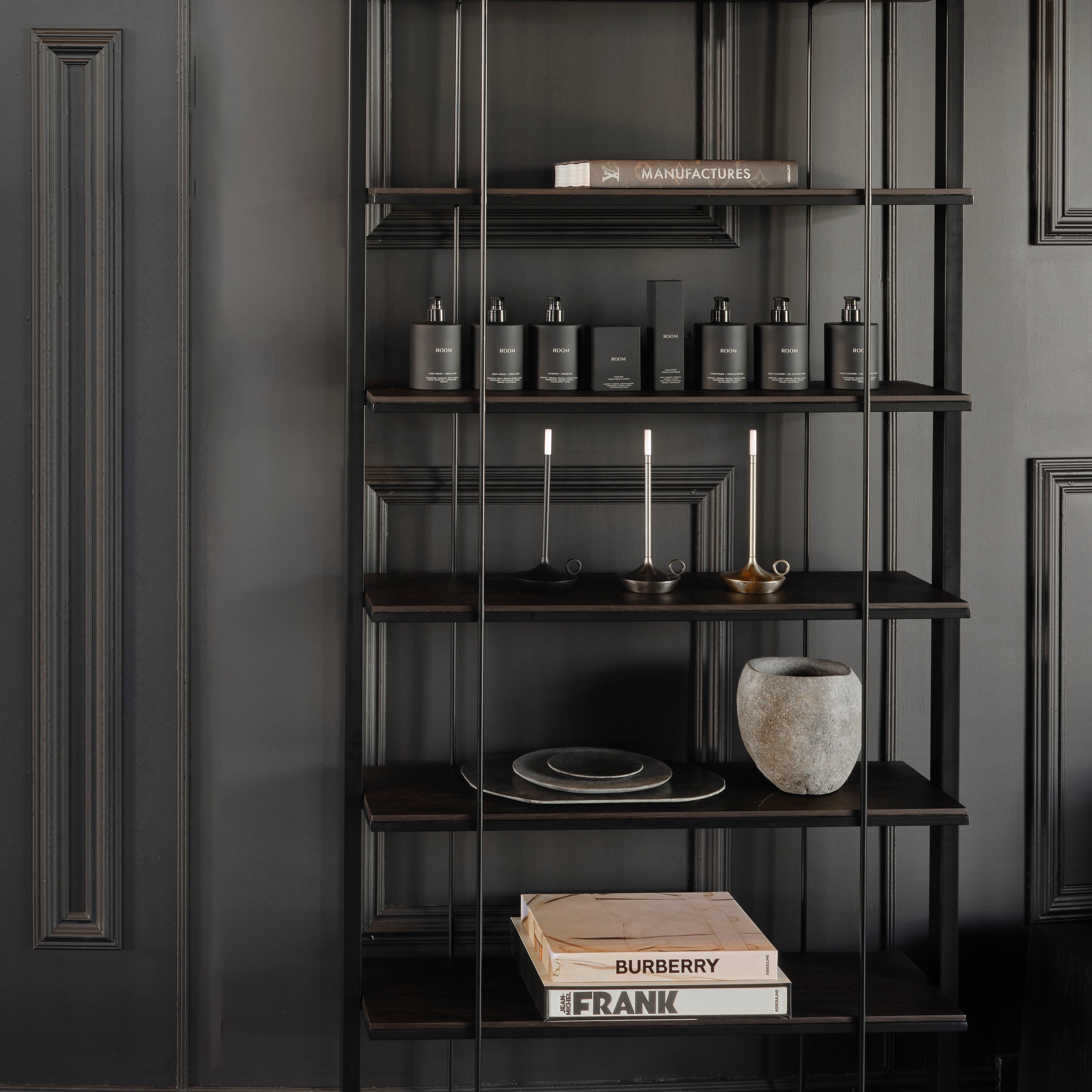 Létagère en bois et métal de Humble Nature, au design moderne en bois foncé, se dresse élégamment contre un mur lambrissé noir. Elle présente des bouteilles noires, trois bougies allumées dans des bougeoirs, un vase gris texturé, une assiette et des livres empilés dont un étiqueté « FRANK ».