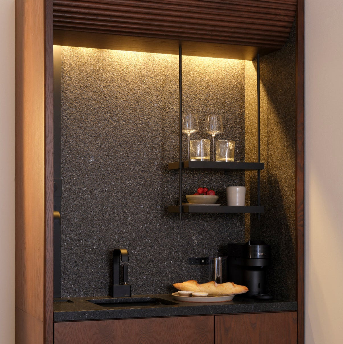 Le BAR CABINET de Humble Nature comprend une kitchenette compacte en bois avec un dosseret en granit, des étagères ouvertes pour les verres, une cafetière, des tasses, des pâtisseries, un éclairage doux et un réfrigérateur intégré, idéal pour une suite Mini Bar élégante.