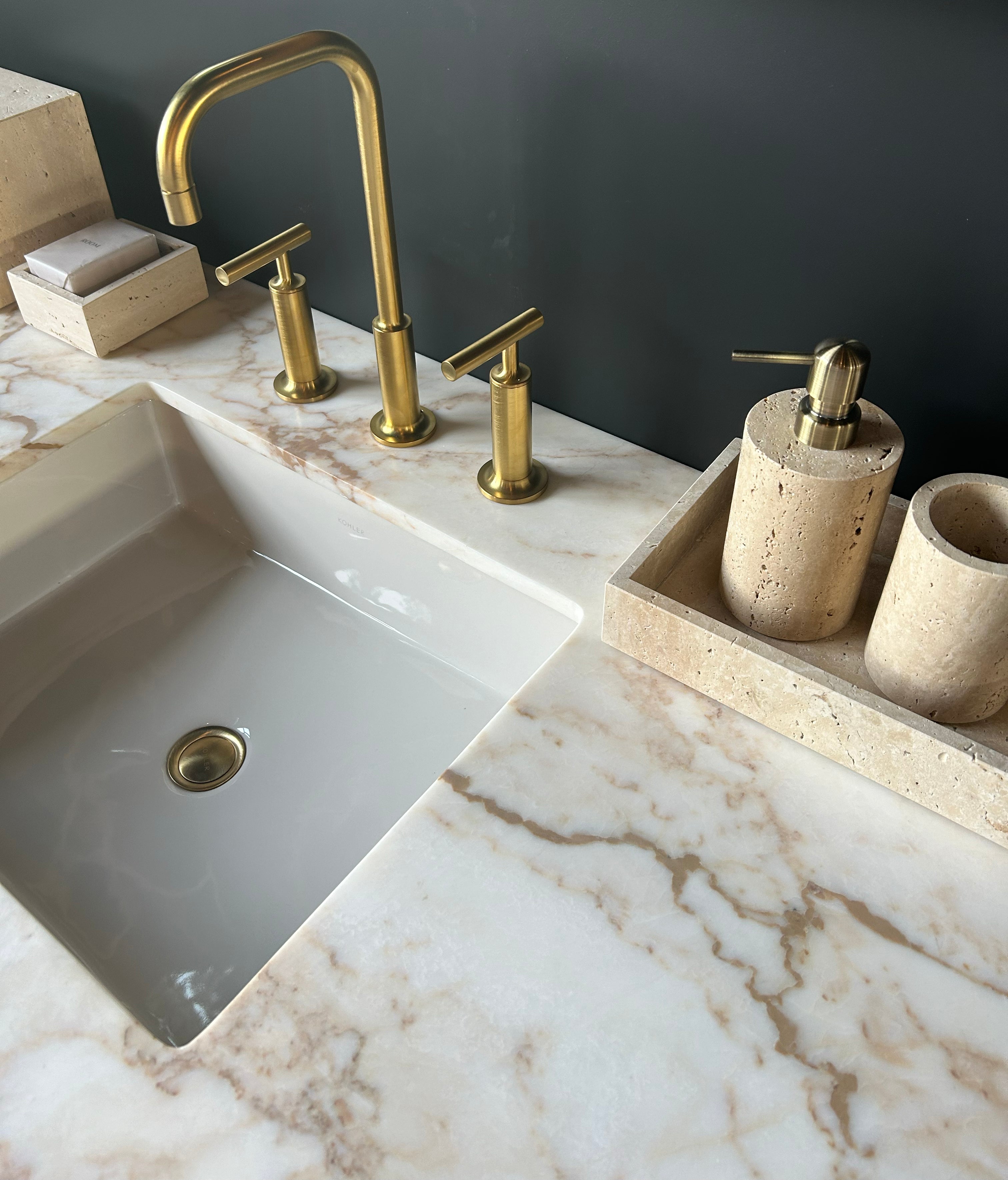 La Vanité Origin | SADDLE de Humble Nature présente un comptoir en marbre avec un évier blanc, un robinet en laiton, un distributeur de savon en pierre beige, un porte-brosse à dents et un porte-savon assorti, le tout sur un mur gris foncé.