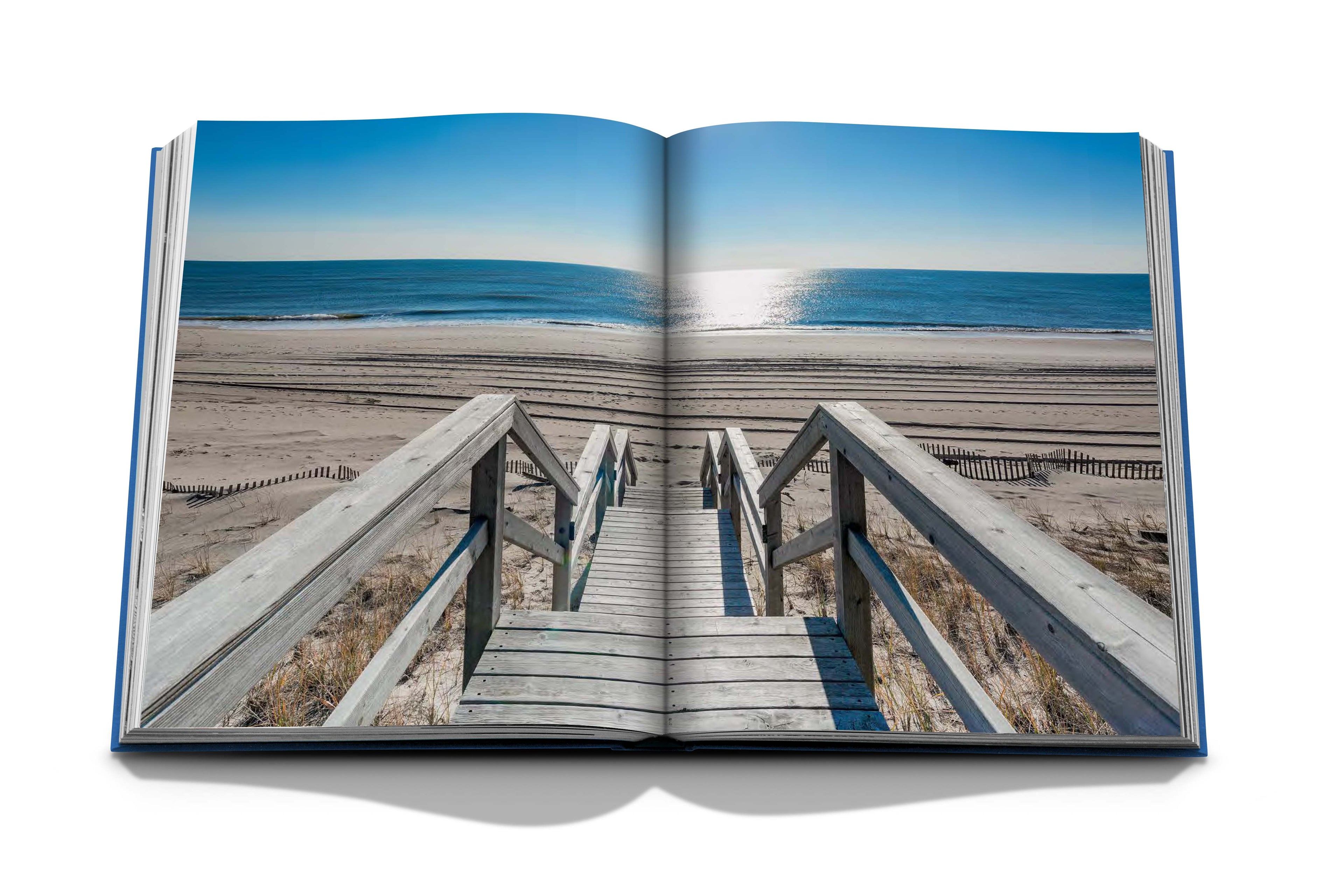 Le livre ouvert « Hamptons Private » dAssouline présente une scène de plage vivante avec des escaliers en bois menant à un rivage sablonneux, des vagues douces sous un ciel bleu clair et la lumière du soleil se reflétant sur locéan, évoquant la paix et le luxe.