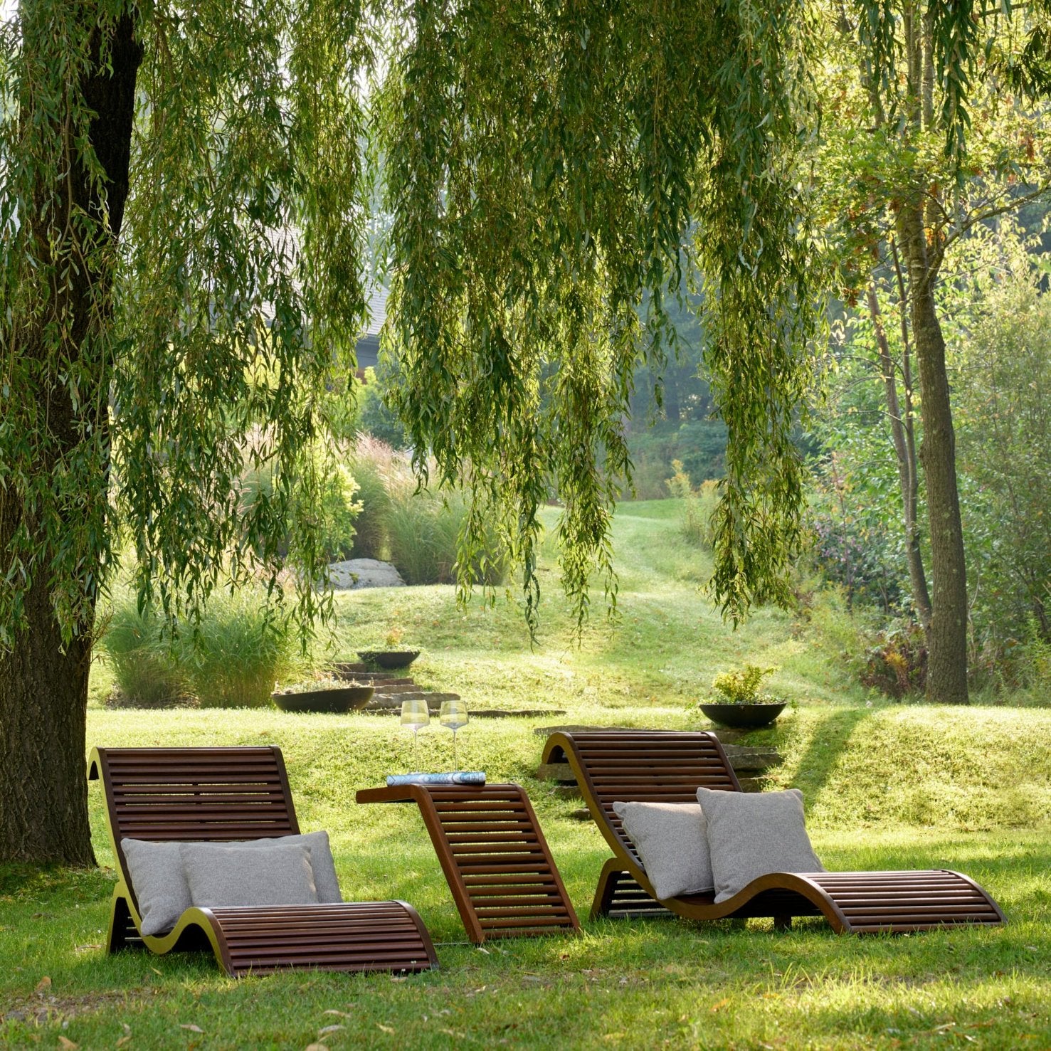 Deux chaises Chaise Lounge Horizon de Humble Nature, avec des coussins gris, sont posées sur une pelouse herbeuse sous un saule. Une petite table en bois accueille un livre et des verres à eau dans le décor dun jardin serein où la nature humble prospère au milieu de la lumière du soleil filtrant à travers les feuilles.