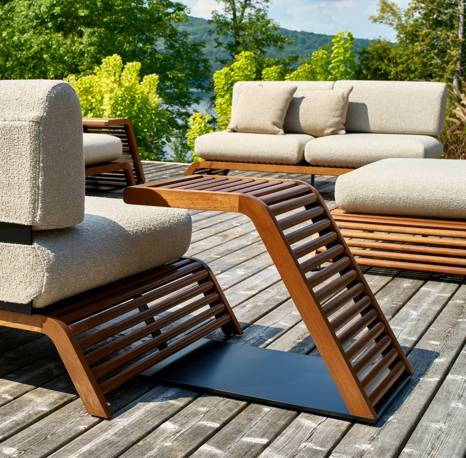 La terrasse extérieure est dotée dun mobilier en rotin moderne, notamment de chaises et de canapés rembourrés de Humble Nature. Le design en lattes de bois de la Table à café Horizon ajoute une touche de qualité supérieure. Des arbres verts luxuriants, un plan deau au loin et une terrasse en bois complètent ce cadre serein.