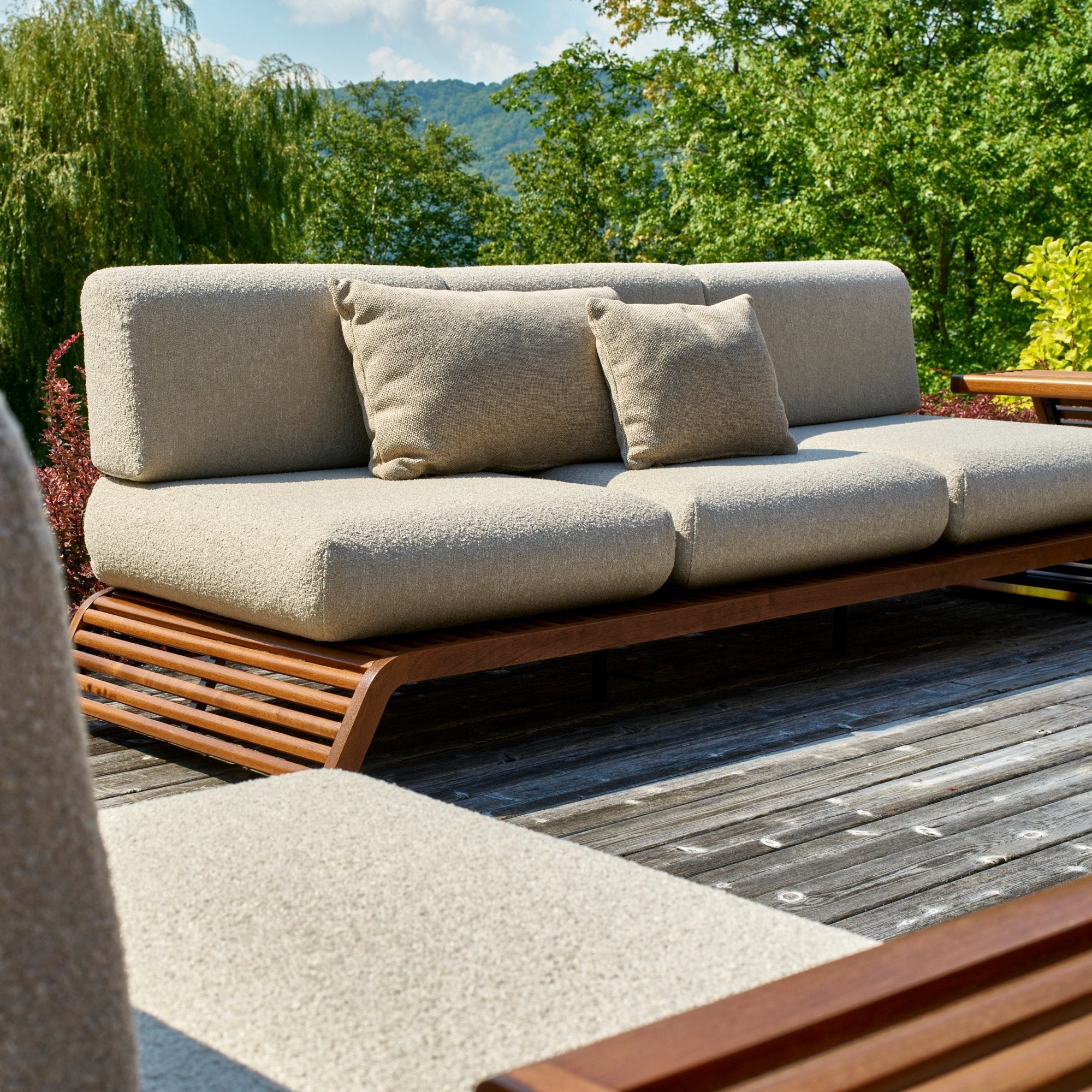 Un patio est équipé du canapé dextérieur triple Horizon de Humble Nature, conçu en beige avec un tissu de haute qualité et deux coussins assortis. Cette pièce élégante repose sur une terrasse en bois au milieu dune végétation luxuriante, darbres et de montagnes lointaines sous un ciel bleu clair.