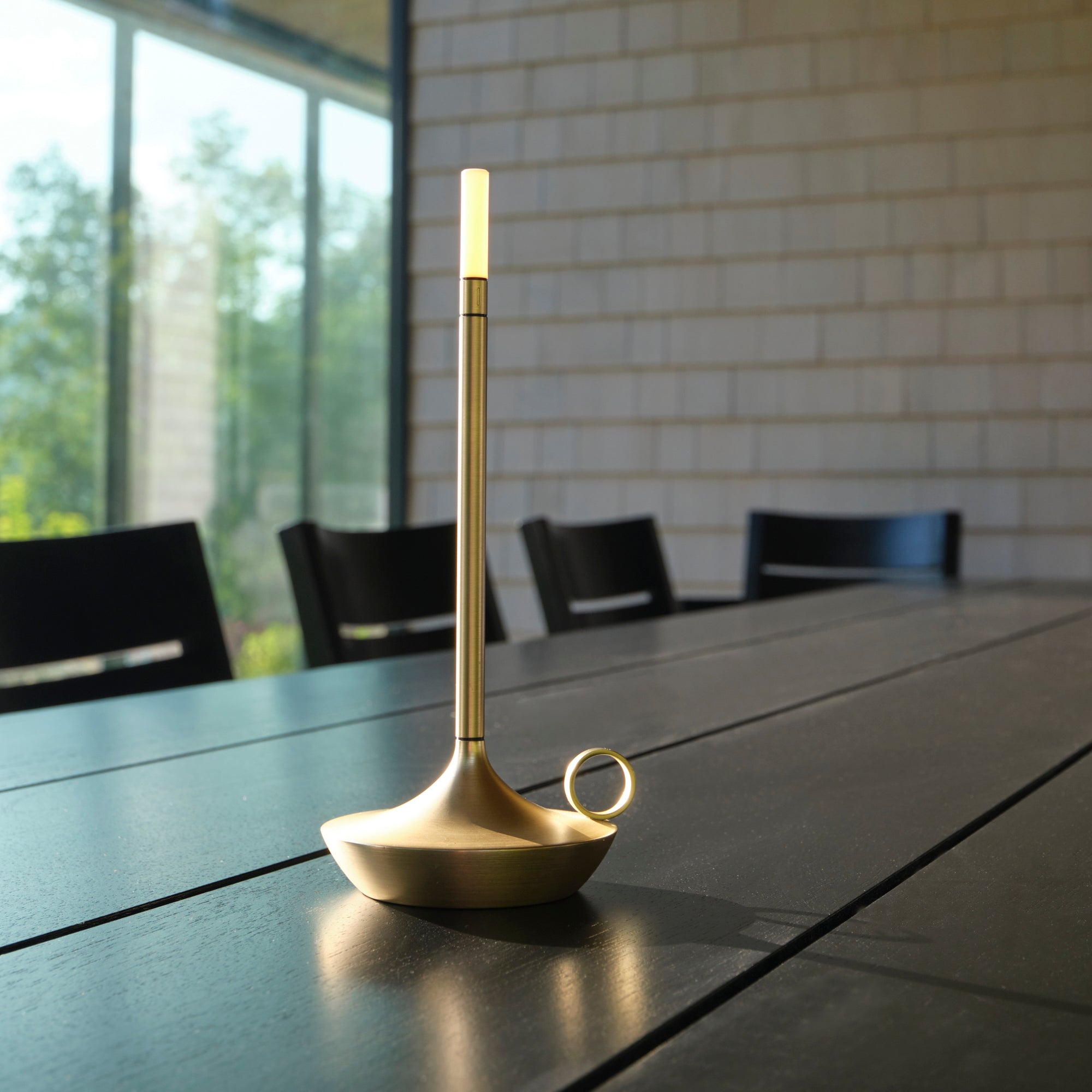 Une Lampe extérieure et intérieure │ SLATE de Humble Nature, moderne et épurée, est posée sur une table en bois foncé, semblable à une lampe à bougie portable. Larrière-plan présente une grande fenêtre avec vue sur les arbres et un mur de briques.