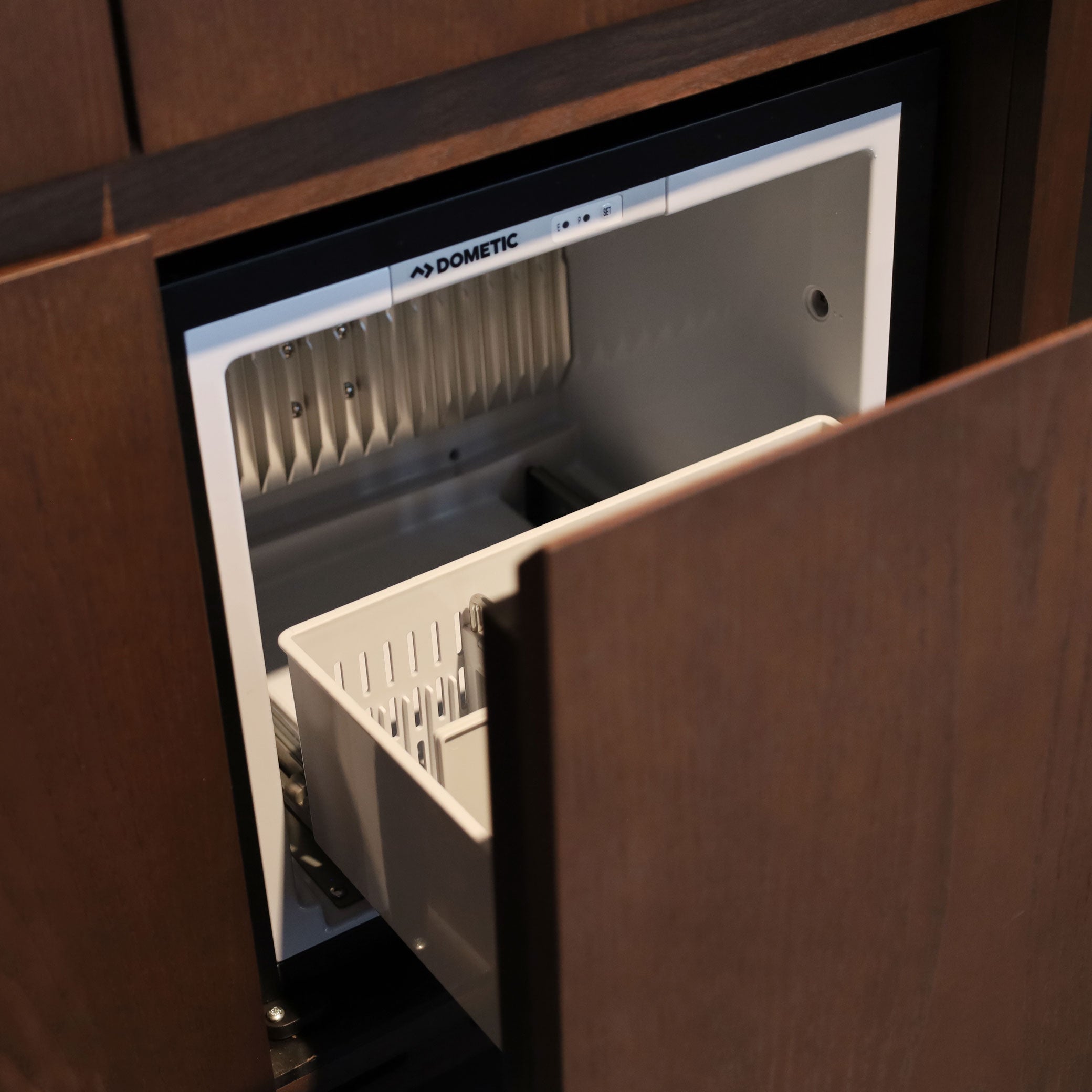 Un BAR CABINET partiellement ouvert de Humble Nature révèle un petit congélateur intégré avec un intérieur blanc et un panier en plastique coulissant derrière des portes en bois, rehaussant le design de luxe de cette suite Mini Bar.