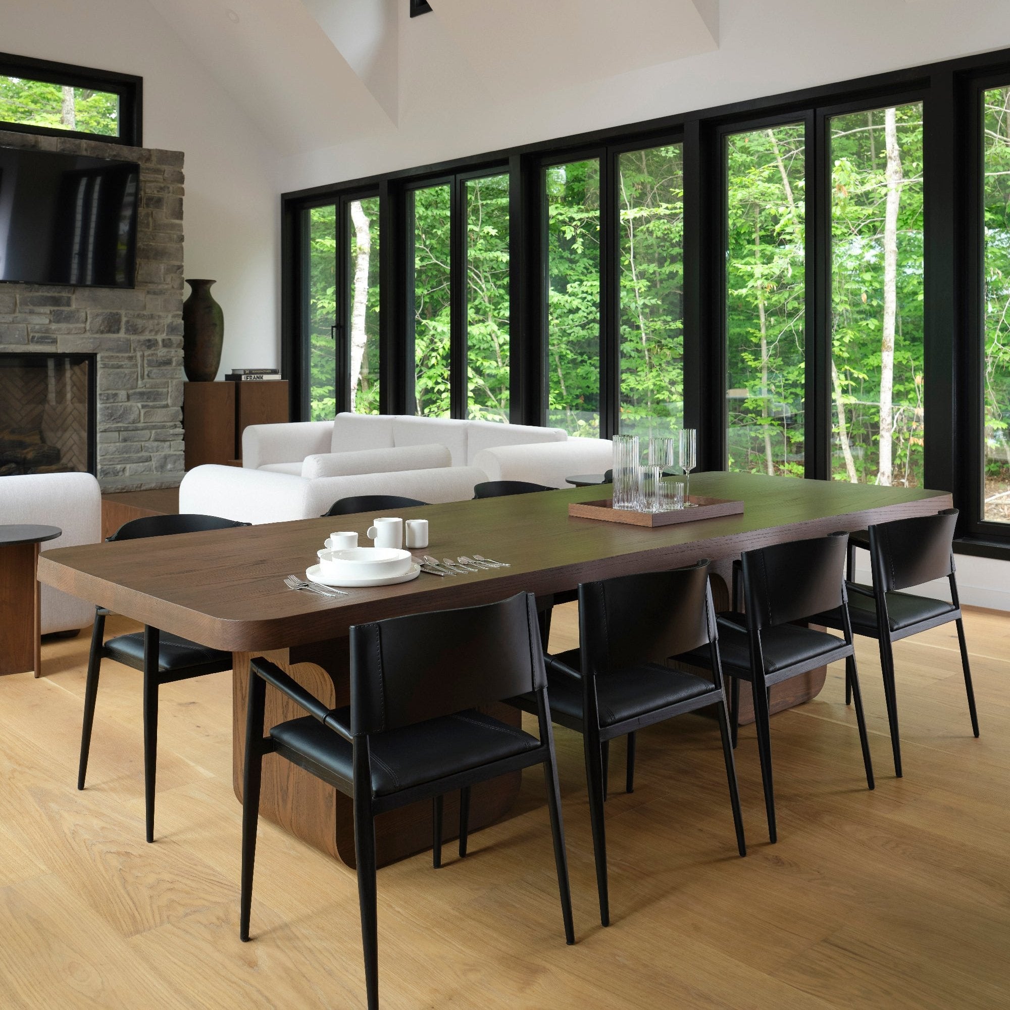 Clin dœil à la nature, la salle à manger moderne est dotée dune longue table en bois et de fauteuils en cuir ONYX de Humble Nature sur un parquet en bois clair. De grandes fenêtres laissent entrevoir une forêt luxuriante, tandis quune cheminée en pierre et des canapés blancs renforcent latmosphère sereine.