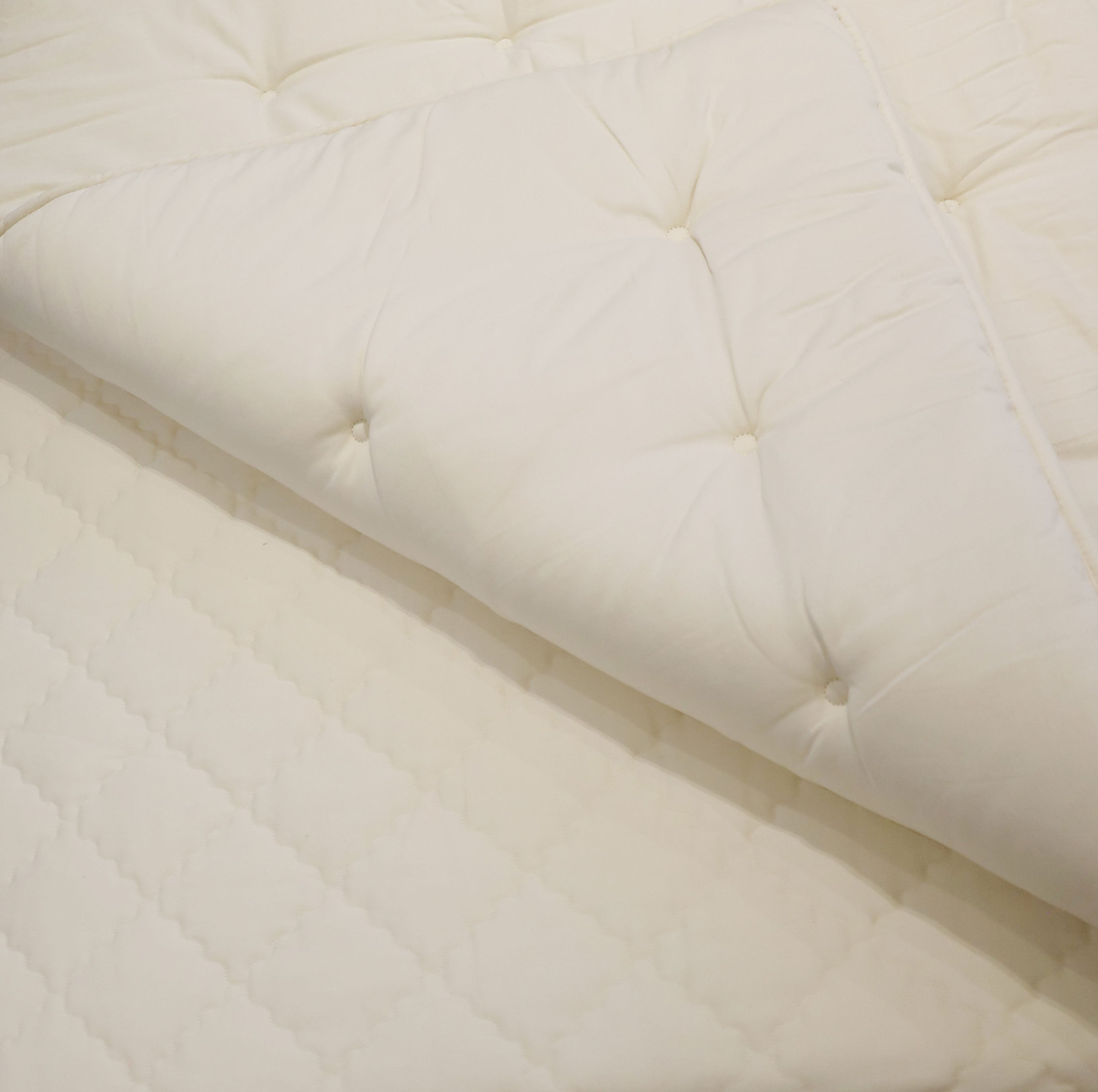Gros plan dun Surmatelas Obasan x Humble Nature │ QUEEN blanc matelassé, présentant un motif en losange avec des boutons touffetés, partiellement plié pour montrer la texture et lépaisseur du rembourrage en dessous pour une amélioration du sommeil.