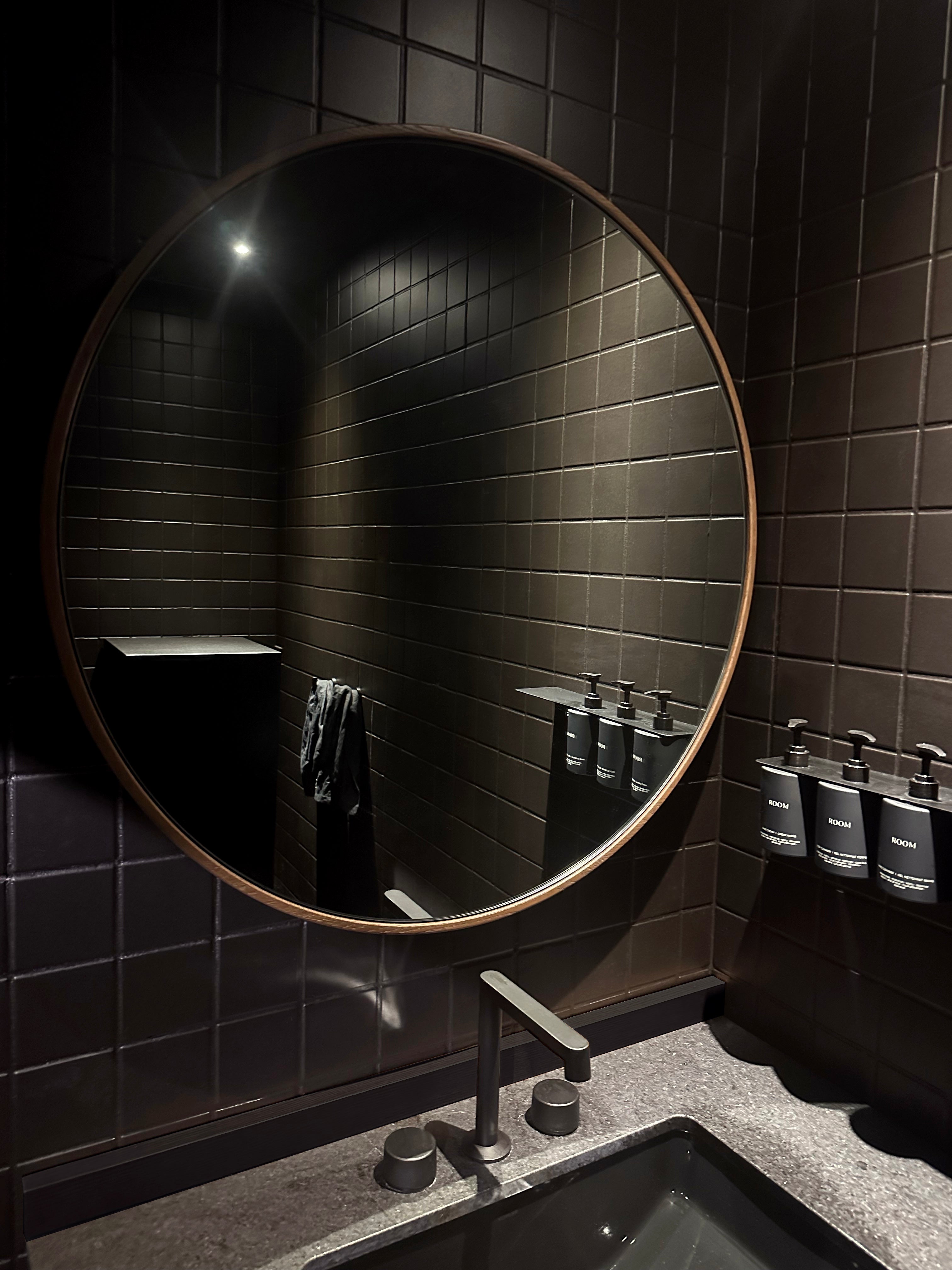 Une salle de bain moderne est dotée de carreaux sombres et dun Miroir 36 rond en chêne rouge Essence de Humble Nature au-dessus dun comptoir gris. Un robinet minimaliste élégant est centré en dessous, dégageant une élégance naturelle. Trois distributeurs muraux avec des étiquettes noires sont alignés à droite.