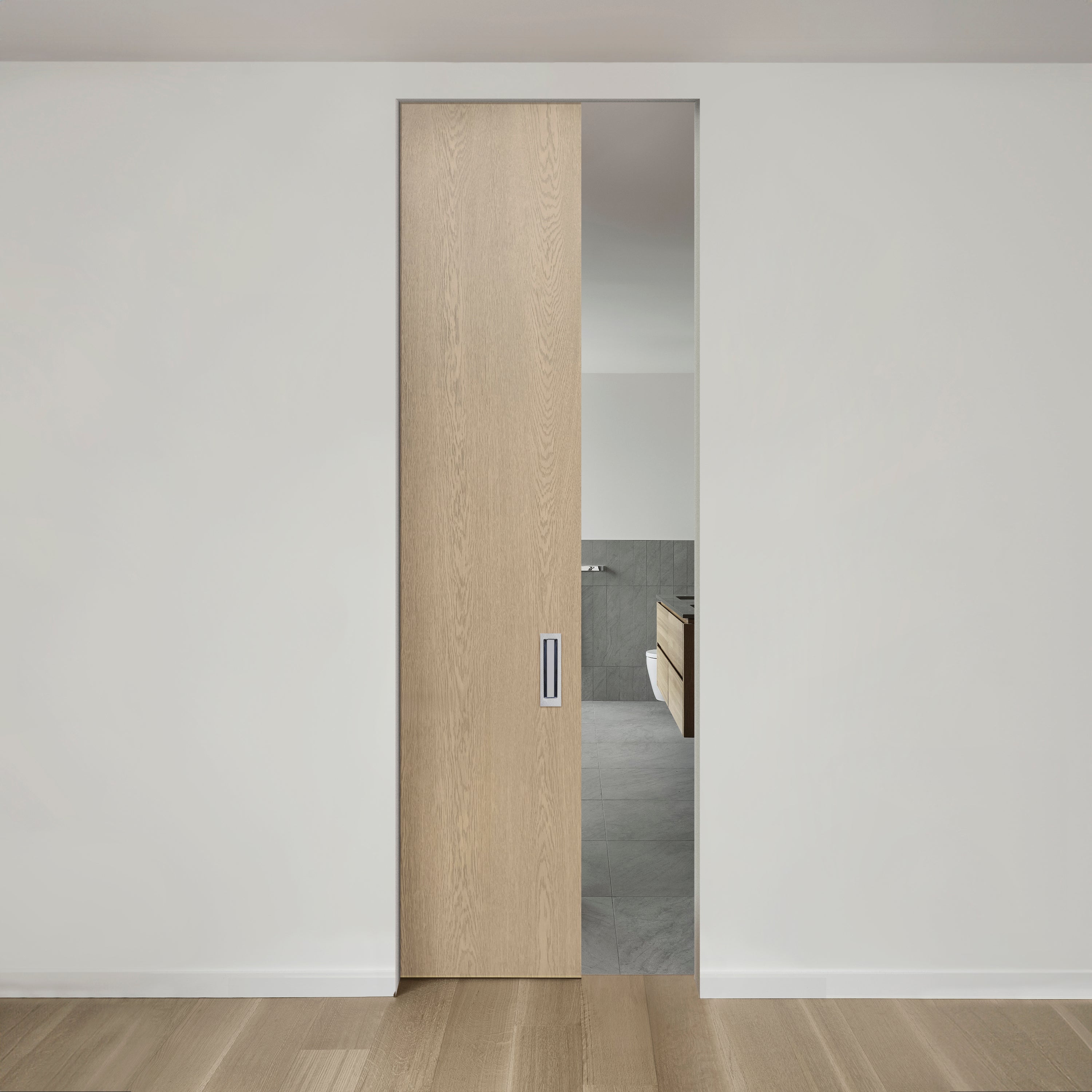 Une porte coulissante en bois minimaliste partiellement ouverte, révélant un aperçu d'une salle de bains moderne avec des carreaux gris et une vanité en bois clair. La pièce présente des tons neutres et épurés et un parquet en bois naturel.