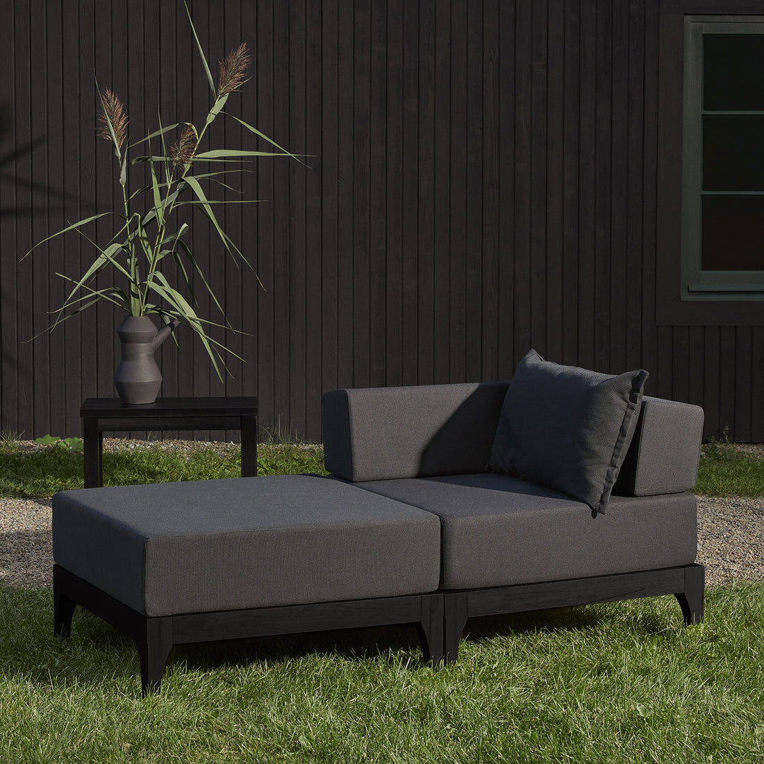 Le canapé dextérieur en bois noir Vivere │ GRAPHITE de Humble Nature repose sur de lherbe verte avec son tissu gris de haute qualité. À côté, une table dappoint assortie avec un vase gris et de hautes herbes complète le look, adossée à un mur en bois foncé avec une petite fenêtre.