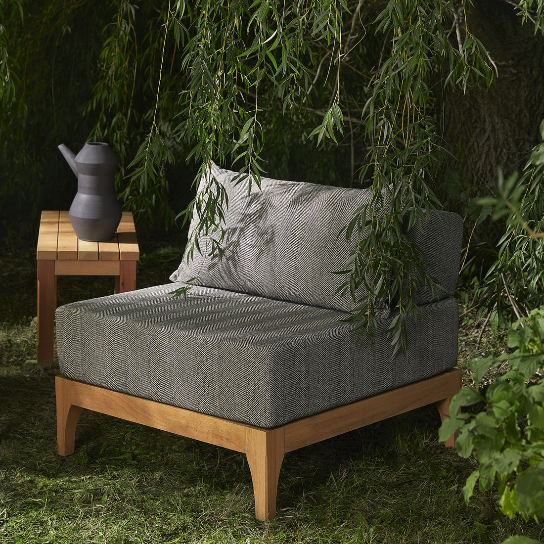 Sous un arbre, entouré de feuilles, le Sofa extérieur en bois naturel Vivere │ NOIR de Humble Nature — une chaise dextérieur grise rembourrée avec des matériaux hypoallergènes et une structure en bois — est associé à une petite table en bois du côté gauche sur laquelle repose un pichet gris foncé. Le décor luxuriant est vert.