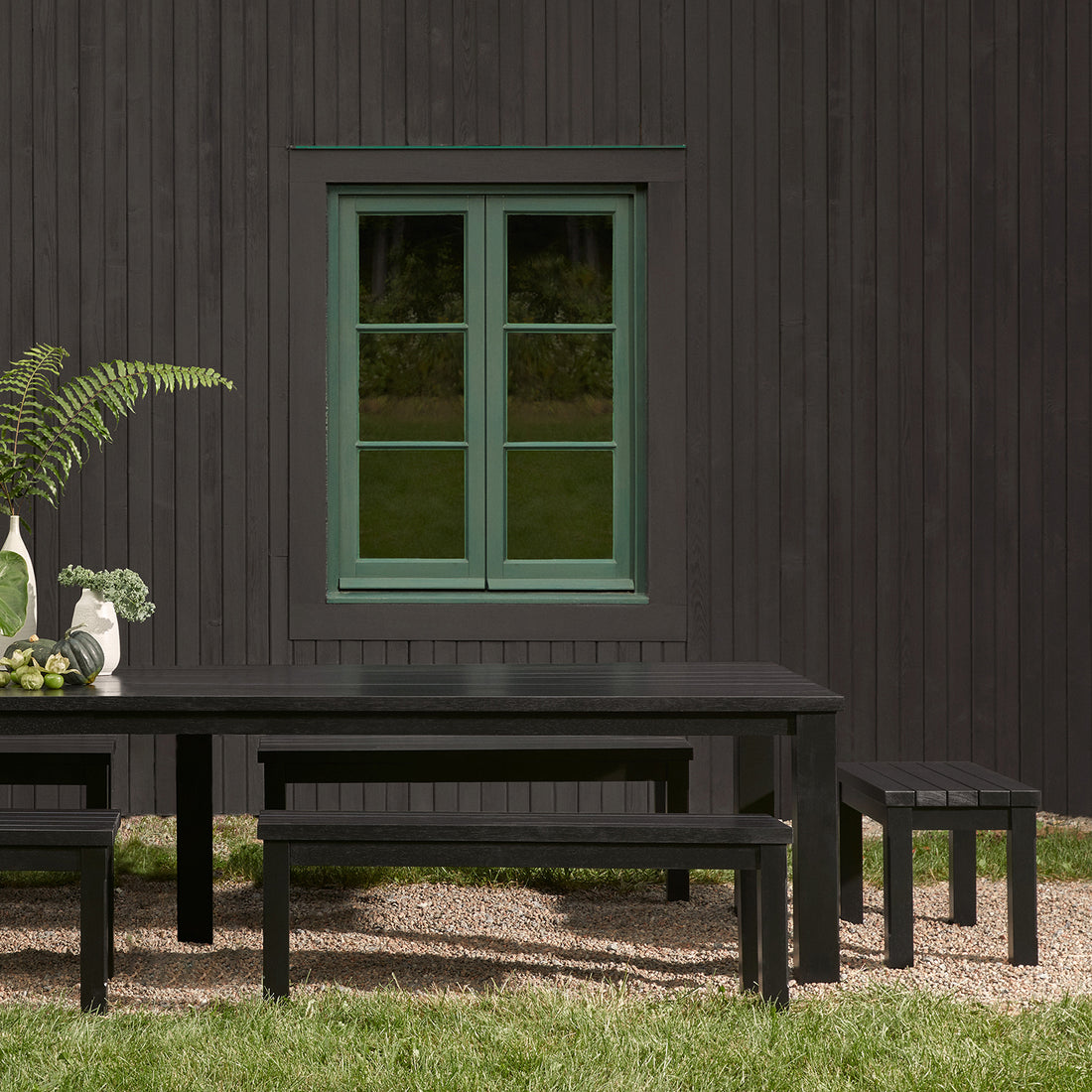 La table extérieure Humble Nature Vivere 66 en Onyx est présentée avec des bancs sur du gravier devant un mur en bois foncé, avec une fenêtre à cadre vert. Un vase avec de la verdure se trouve au sommet de la table, complétant le paysage tranquille de lherbe et des arbres.