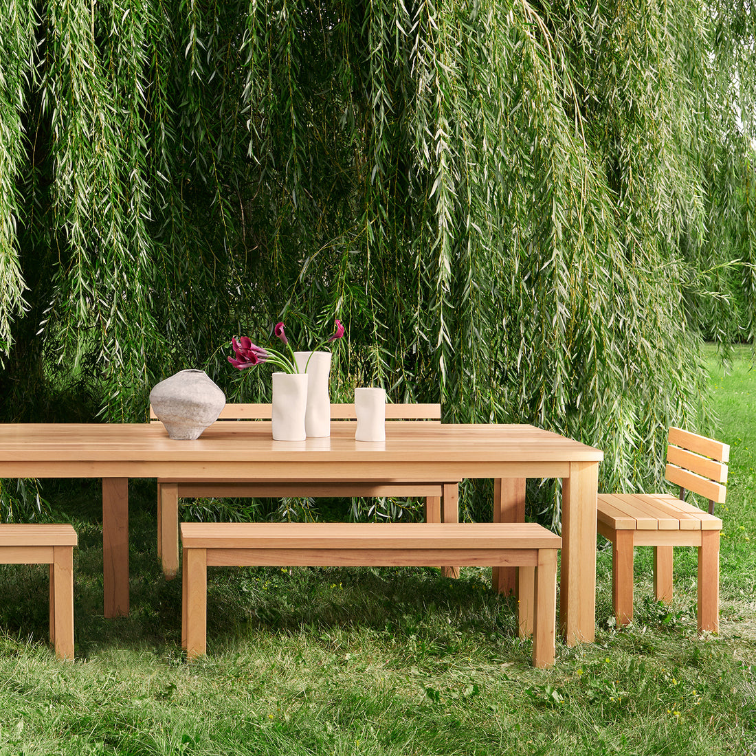 Sous un saule pleureur luxuriant, une configuration de salle à manger extérieure sereine de Humble Nature comprend la Table extérieure Vivere 132 │ Naturel avec des bancs et une chaise. Faisant partie de nos collections de table écoresponsables, elle est ornée de vases blancs contenant des fleurs violettes et dun vase rond en forme de pierre sur de lherbe verte.