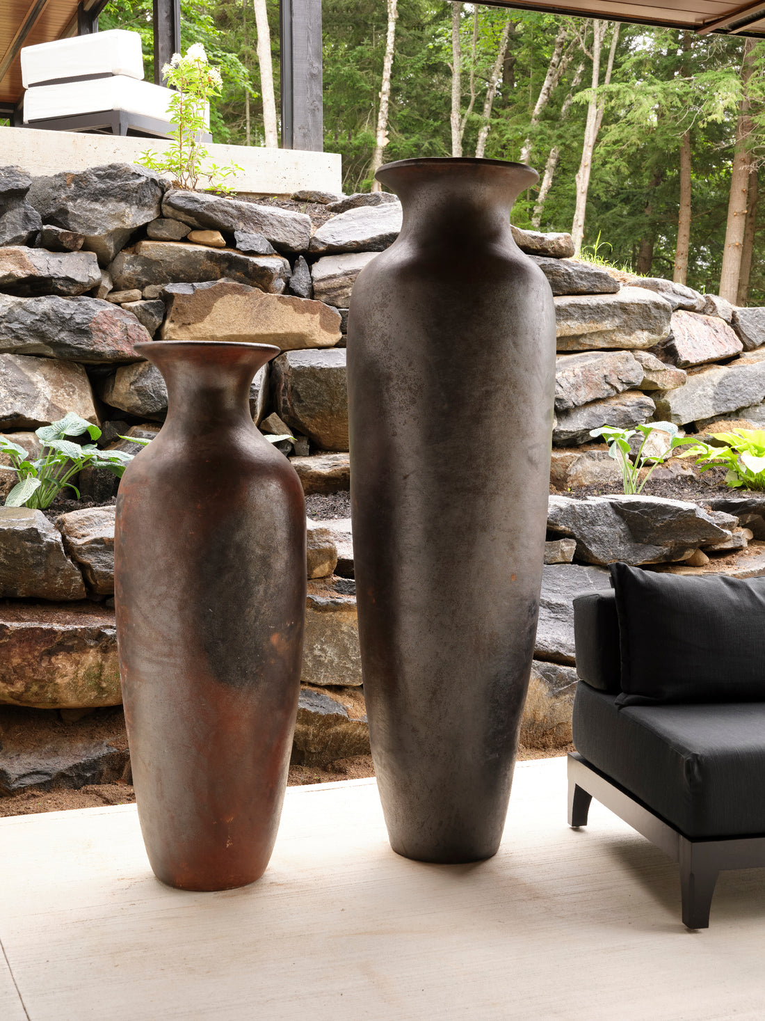 Deux grands vases Pot en argile de Humble Nature de différentes tailles sont disposés sur une terrasse à côté dun canapé dextérieur noir, fabriqué avec un savoir-faire traditionnel. Ils complètent les murs en pierre empilés et le décor de verdure luxuriante.