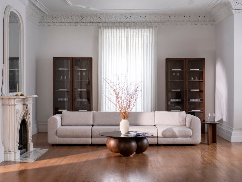 Un salon spacieux est doté dun canapé modulable beige clair de la collection Suite Sofa de Humble Nature, face à une cheminée en marbre. Une table basse ronde avec des branches sur le plancher en bois ajoute du charme, tandis que deux vitrines de luxe encadrent la grande fenêtre pour un design élégant.