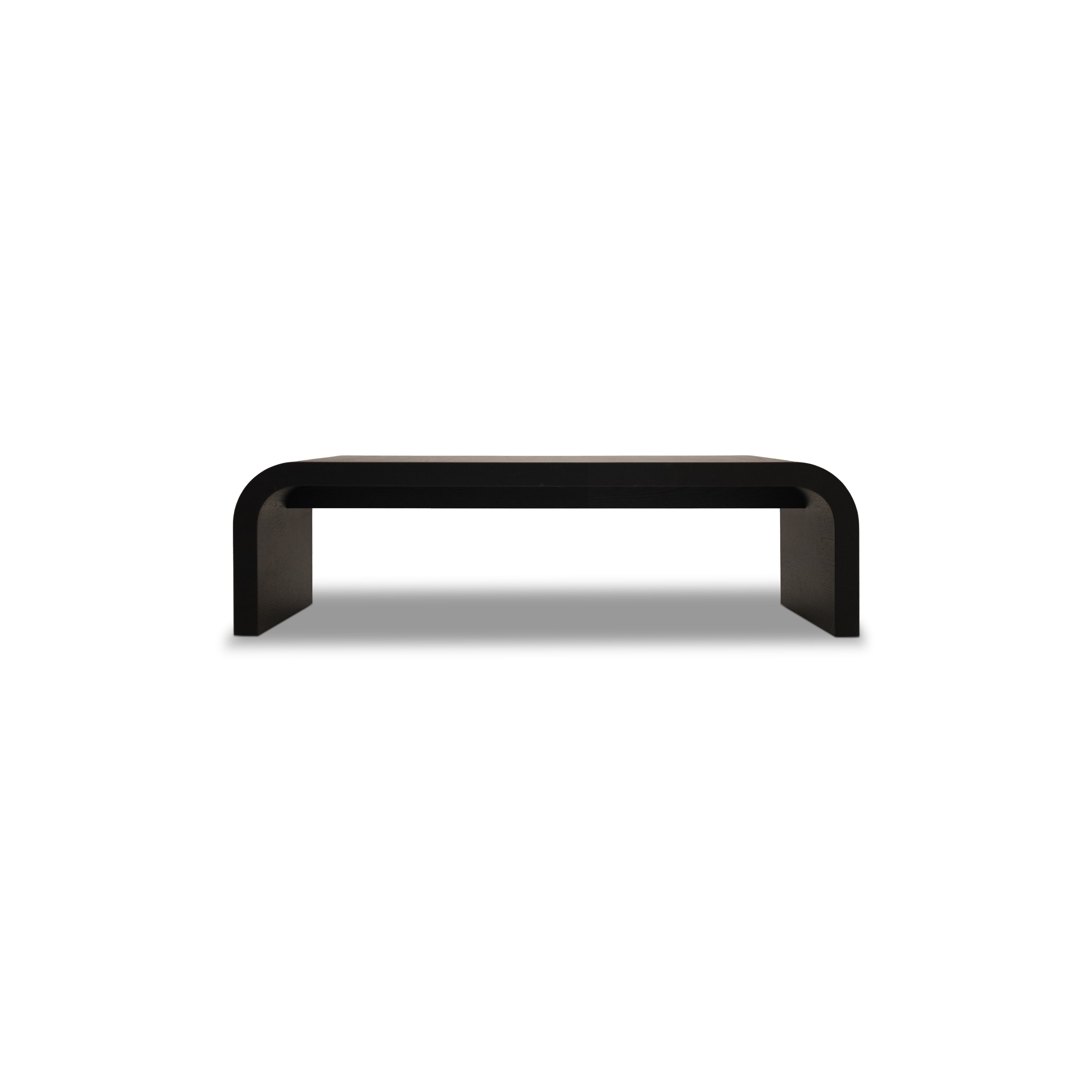 Le Banc 64 en bois Essence │ ONYX de Humble Nature est une banc noir minimaliste avec une surface rectangulaire lisse et des bords élégamment incurvés, reflétant la sophistication alors quelle se dresse gracieusement sur un fond gris clair.