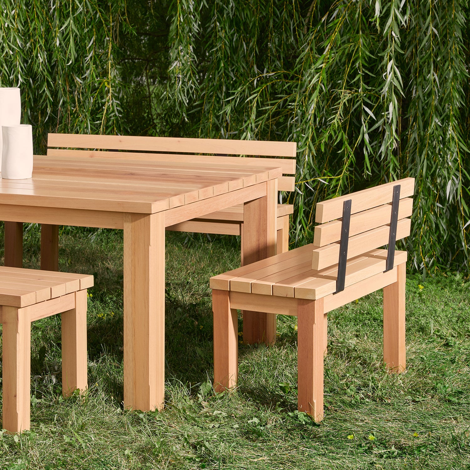 Le Banc extérieur avec dossier Vivere de Humble Nature est un ensemble de table dextérieur en bois avec bancs et chaises écologiques, niché sur une pelouse herbeuse. De grands arbres feuillus offrent un décor tranquille, idéal pour les réunions ou les repas en plein air.