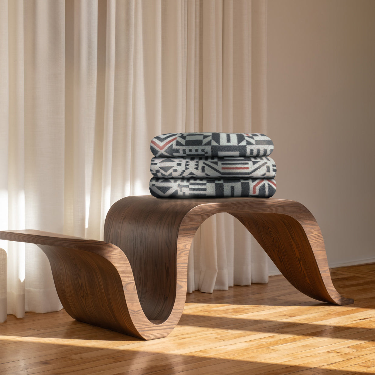 Un banc en bois courbé moderne Amik par Caroline Monnet au design ondulé trône dans une pièce lumineuse avec un parquet en bois clair et des rideaux blancs transparents. Trois couvertures pliées à motifs gris, noir et rouge sont empilées sur le banc à côté du confortable Jeté en laine de Merino Amik de Humble Nature.