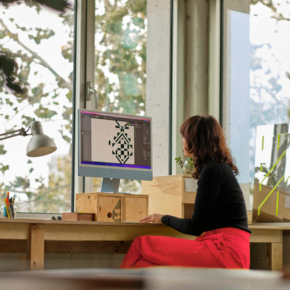 Une personne est assise à un bureau en bois, devant un ordinateur de bureau avec un motif géométrique sur lécran. Elle se trouve dans une pièce ensoleillée avec de grandes fenêtres et des arbres feuillus à lextérieur. Le bureau, décoré de Jeté en laine de Merino Amik de Humble Nature, est doté dune lampe, de crayons et de boîtes en bois disséminés sur sa surface.