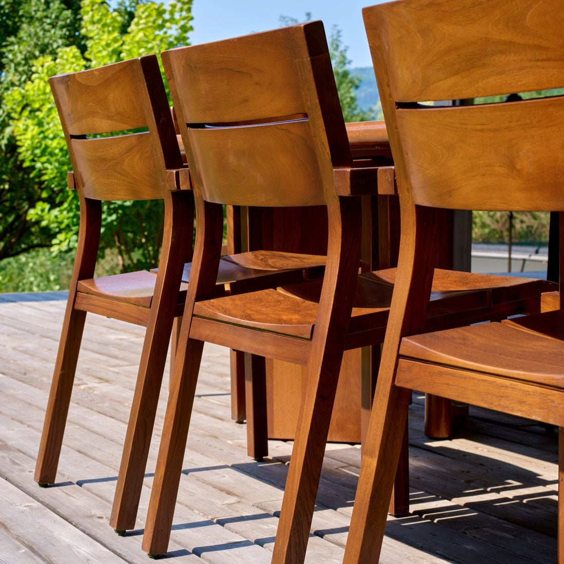 Les Chaises extérieures en bois de teck │ Roussette de Humble Nature sont disposées sur une terrasse ensoleillée, entourée dune verdure luxuriante et dun ciel bleu clair.