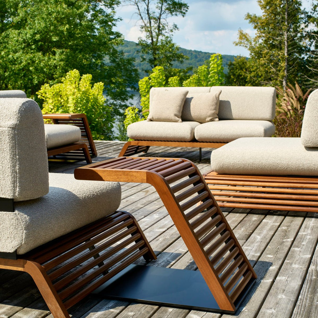 Le canapé double extérieur Horizon en crème bouclée de Humble Nature est doté de sièges rembourrés et de cadres en bois hypoallergéniques, installés sur une terrasse avec des arbres luxuriants et une vue sur leau sous un ciel partiellement nuageux.