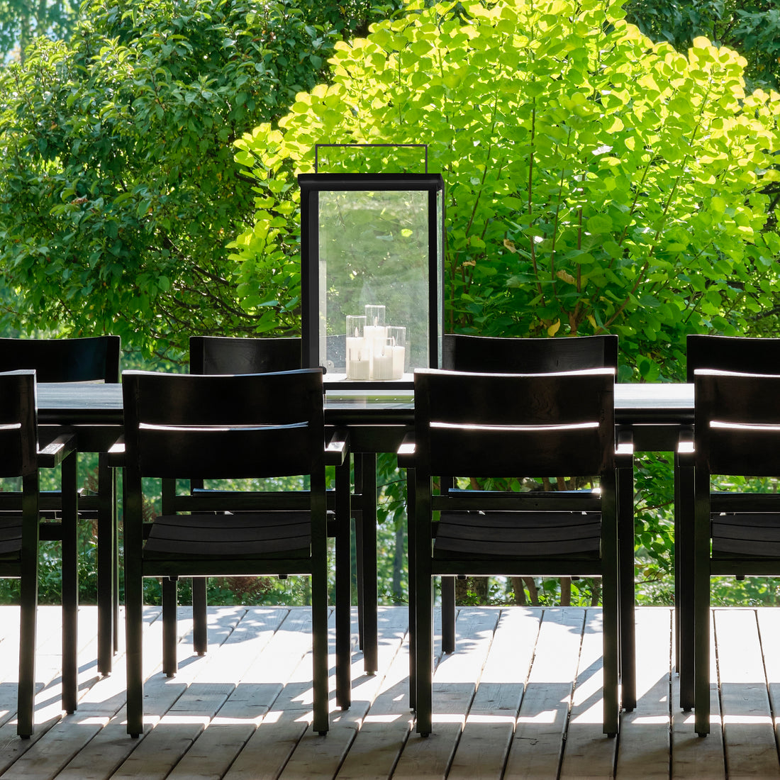 La salle à manger extérieure comprend une chaise extérieure en bois de teck noire de Humble Nature disposée autour dune table sur une terrasse en bois écologique. Une grande lanterne avec des bougies orne la table tandis que des arbres verts luxuriants et une lumière du soleil éclatante en arrière-plan renforcent latmosphère sereine.