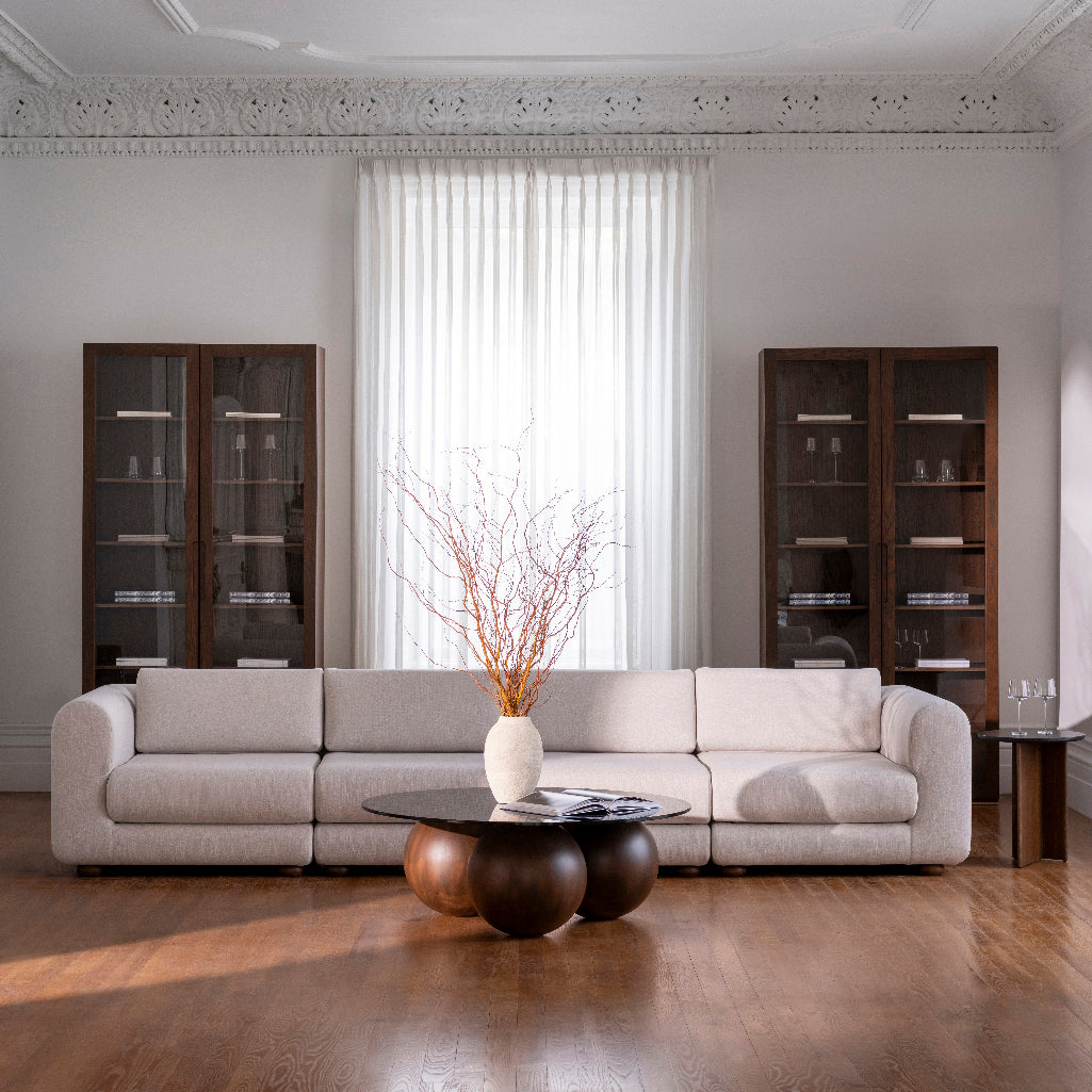 Un salon moderne présente la suite Humble Nature Sofa Simple en beige avec un tissu luxueux, une table basse ronde avec trois sphères marron comme base et un grand vase avec des branches. Deux armoires en bois abritent la décoration à côté dune grande fenêtre avec des rideaux transparents derrière le canapé.