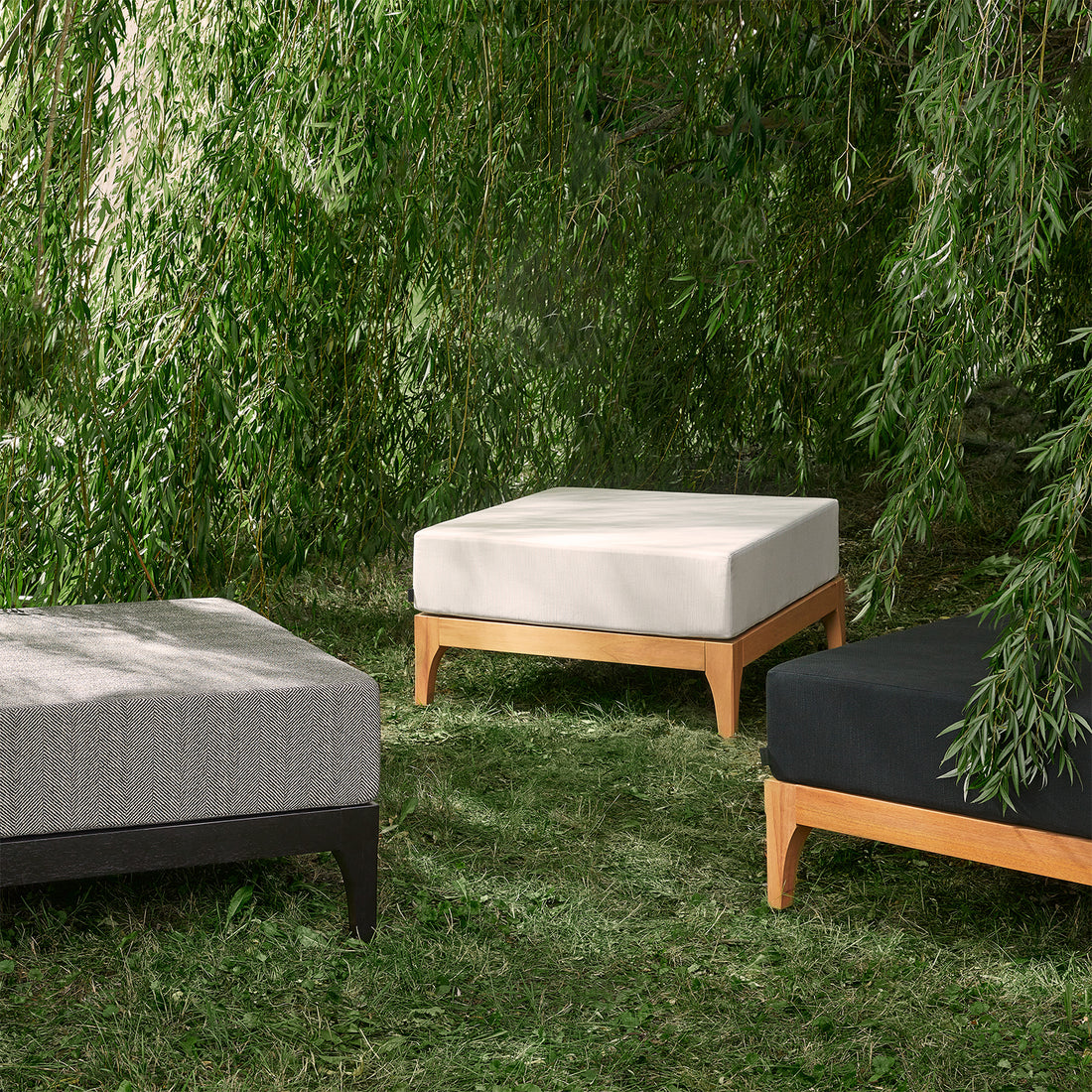 Trois poufs dextérieur en bois noir Vivere de Humble Nature avec des plateaux rembourrés en tissus de haute qualité sont disposés sur lherbe sous des branches vertes luxuriantes en surplomb, dans des tons de gris clair, beige et gris foncé. Cela crée une atmosphère sereine et naturelle.