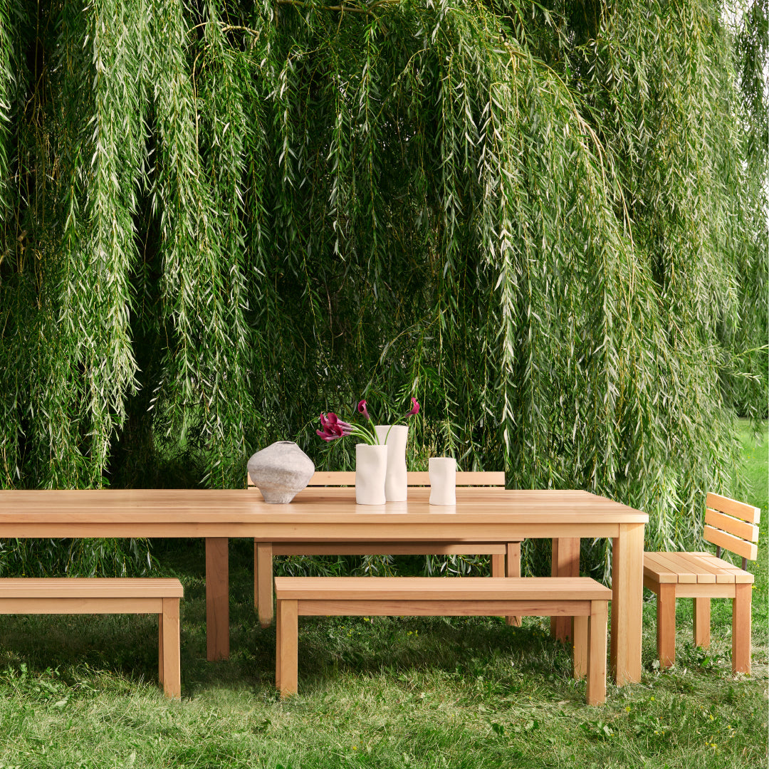 Laménagement extérieur Humble Nature comprend la table et les bancs en bois Banc long extérieur Vivere │ Naturel sous un saule luxuriant, orné de vases blancs de fleurs violettes et dun ornement en pierre grise, entouré dherbe.