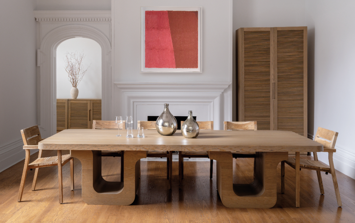 Salle à manger élégante et minimaliste avec une grande table en bois au design sculptural, entourée de chaises assorties en bois clair. En arrière-plan, une cheminée blanche ornée d’un tableau abstrait rouge et des armoires en bois avec des lignes verticales. Décor apaisant aux tons naturels, mettant en valeur des éléments de design contemporain et chaleureux.