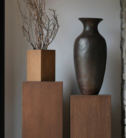 Dans un intérieur minimaliste, un grand vase en céramique foncé est exposé sur la Table piédestal carré 41 Essence | Saddle de Humble Nature, positionné à côté d&