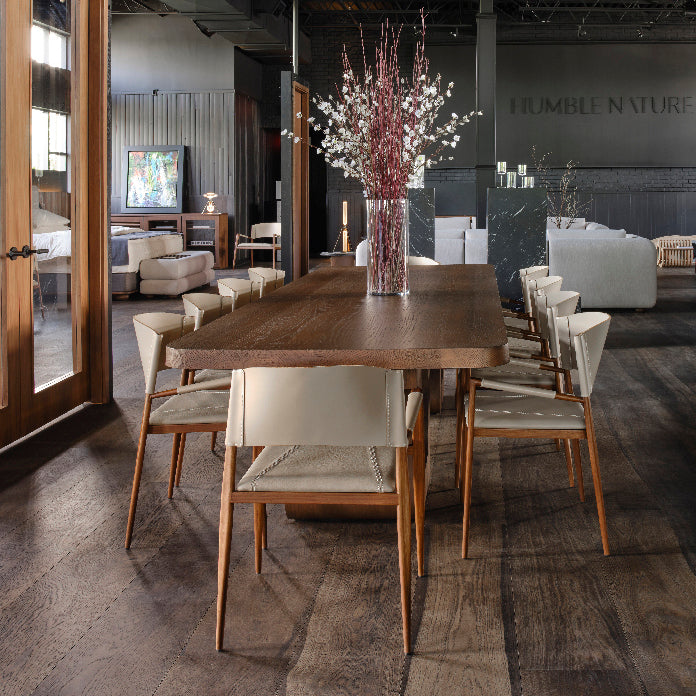 Une salle à manger moderne présente une longue table en bois avec huit chaises blanches à dîner avec accoudoirs Suite de NUTSHELL. Un grand vase aux branches rouges et blanches constitue le centre de lexposition. En arrière-plan, des canapés moelleux complètent les œuvres dart de Humble Nature, créant une ambiance de salon chaleureuse.