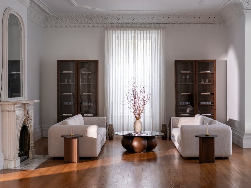 Le salon est composé de deux canapés beiges qui se font face, avec une table basse ronde au milieu, ornée de branches décoratives. De chaque côté de la fenêtre se trouvent deux armoires Humble Nature Cabinet à double finition Pecan | Porte verre, et à gauche se trouve une cheminée ornée de rideaux transparents.