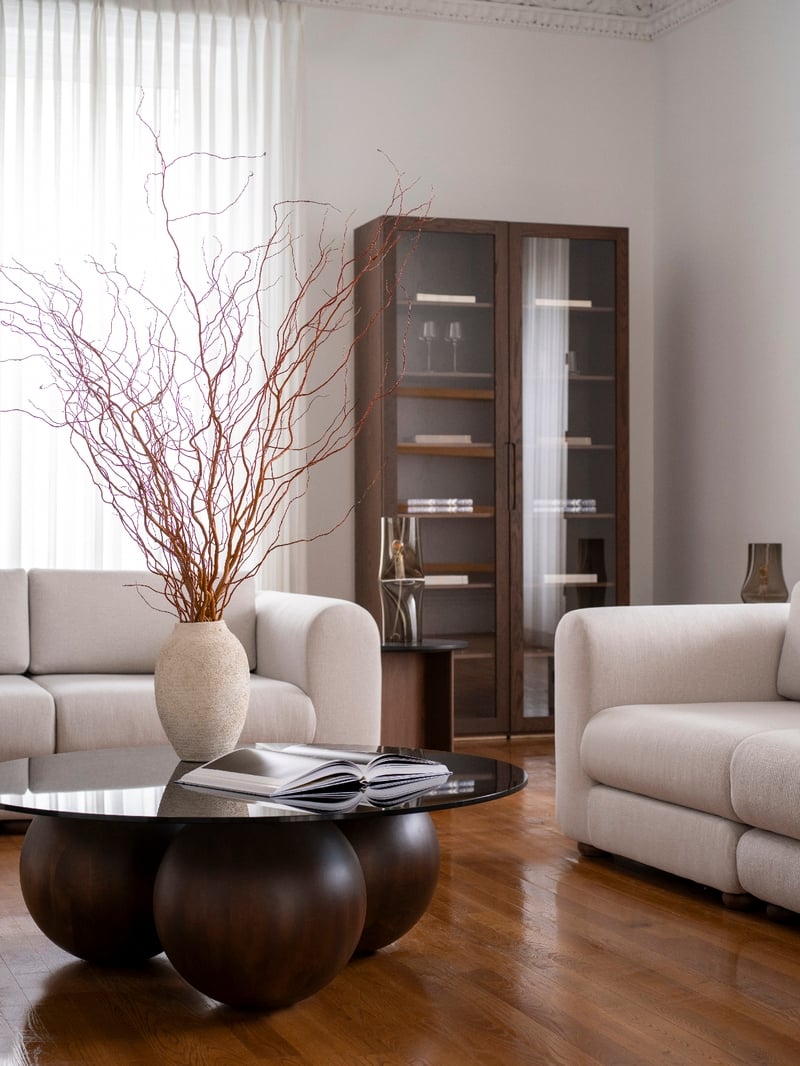 Un salon moderne est doté de la suite Sofa coin droit de Humble Nature, avec ses élégants sièges blancs. Une table basse à base sphérique accueille un livre et un vase de branches torsadées. Une grande armoire en verre se trouve à larrière tandis que des rideaux transparents illuminent le luxueux plancher en bois.