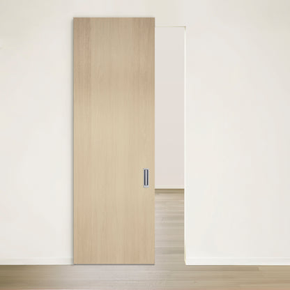 Une pièce minimaliste et blanche avec un sol en bois clair est dotée dune simple porte coulissante en bois clair légèrement ouverte. Cette porte est la « Porte Pure coulissante en chêne blanc placage brossé sur dosse » de Humble Nature et comprend une poignée rectangulaire moderne sur le côté droit.