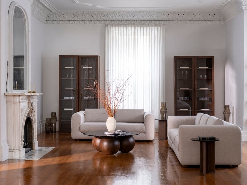 Salon élégant avec une suite ottomane de Humble Nature avec des canapés beiges, une table basse ronde en bois et un vase blanc avec des branches. Deux armoires hautes encadrent une fenêtre à rideaux, tandis quune cheminée décorative avec un grand miroir rehausse le design sophistiqué.